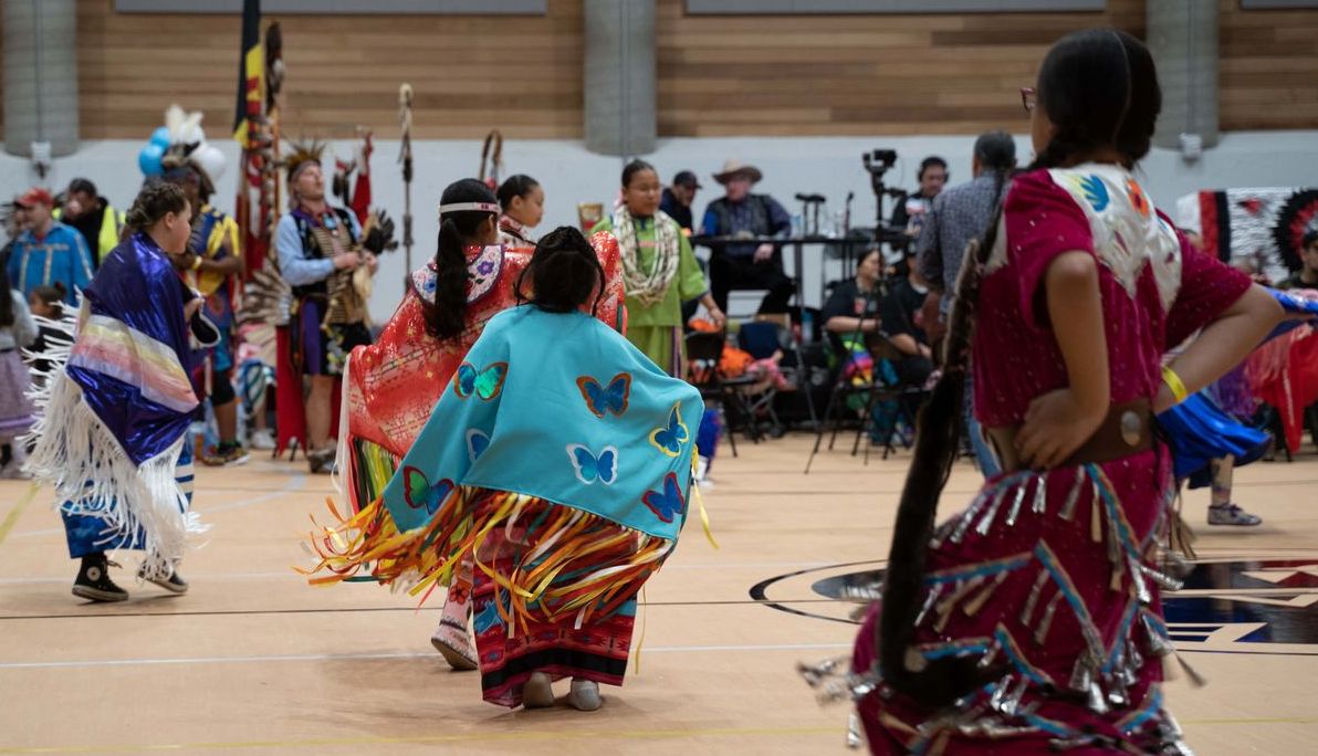 Youth wearing fancy dress shawls and jingle dresses dance in a gym space.